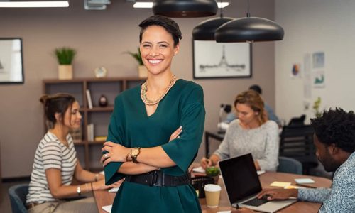 Portrait of successful business woman standing with her colleagues working in background at office. Portrait of cheerful fashion girl in green dress standing with folded arms and looking at camera. Beautiful businesswoman feeling proud and smiling.
